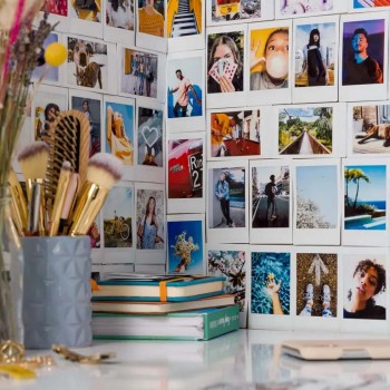 Fujifilm Instax Mini Film-collagevägg och tillbehör. Flera färgglada polaroidbilder på väggen som en collage, framför makeupborstar i en grå burk och böcker i en hög.